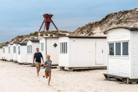 Dreng og mand løber på stranden foran hvide hytter med sandklitter og et rødligt vartegn i baggrunden.