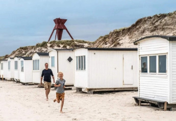 Un garçon et un homme courent sur la plage devant des cabanes blanches, dunes et structure rouge en arrière-plan.
