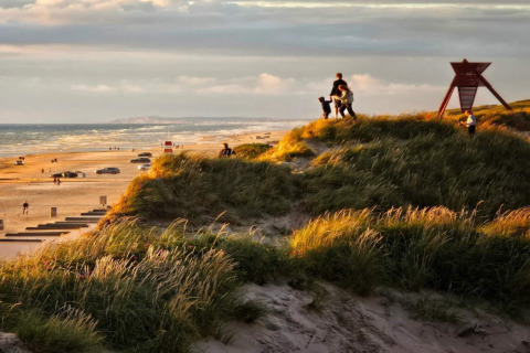 Solnedgang ved klitterne på en dansk strand med familier, biler og udsigt over havet fra en lodge.