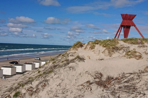 Sanddünen und rote Landmarke an einem sonnigen Strand in Blokhus, Dänemark, mit weißen Hütten.