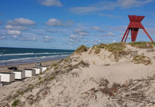 Sandklitter, hytter og et rødt vartegn ved stranden i Blokhus, Danmark, på en solrig dag med blå himmel.