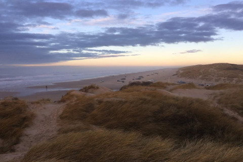 Udsigt fra en hytte over klitterne ved stranden med flere biler parkeret langs kystlinjen ved solnedgang.