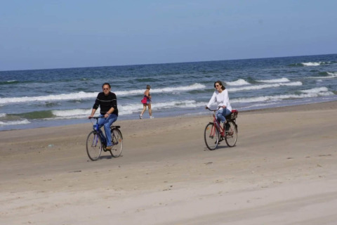 To personer cykler på stranden nær havet, mens en person går i baggrunden på en solrig dag.