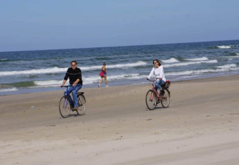 To personer cykler på stranden nær havet, mens en person går i baggrunden på en solrig dag.
