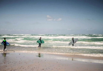 Tre personer med surfbrætter går ud i havet ved en strand nær en lodge under en lys himmel.