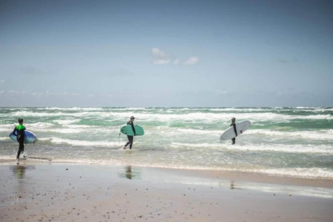 Drie surfers met surfplanken lopen de zee in vanaf een strand nabij een lodge onder een heldere lucht.