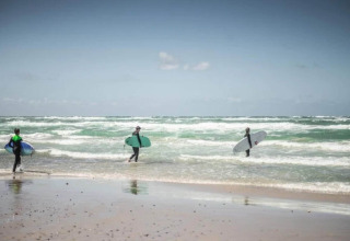 Drie surfers met planken gaan het water in aan een strand bij een lodge onder een open hemel.
