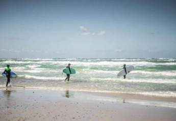 Tre surfisti con tavole camminano verso il mare su una spiaggia vicino a un lodge sotto un cielo limpido.