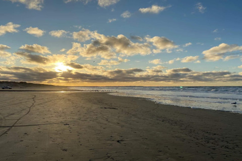 Sonnenuntergang am Strand bei einer Lodge, mit Wellen, Sand und lebhaften Wolken am Himmel.