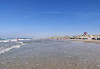 Zonnig strand met mensen, geparkeerde auto's en duinen op de achtergrond onder een heldere blauwe lucht.