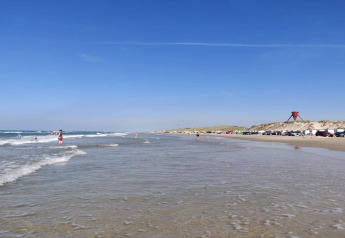 Zonnig strand met mensen, geparkeerde auto's en duinen op de achtergrond onder een heldere blauwe lucht.