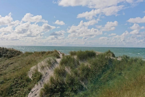 Uitzicht vanuit een lodge over grasrijke zandduinen en de zee, onder een licht bewolkte blauwe lucht.