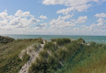 Uitzicht vanuit een lodge over grasrijke zandduinen en de zee, onder een licht bewolkte blauwe lucht.