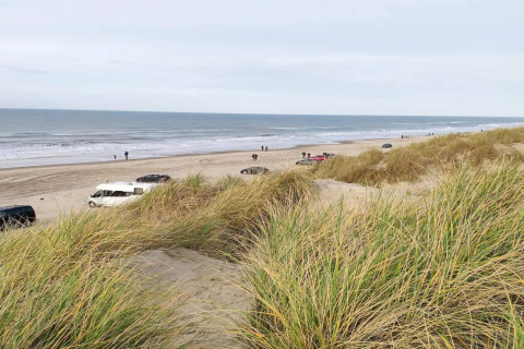 Uitzicht vanuit een lodge op een strand met auto's, grasduinen en mensen langs de zee.