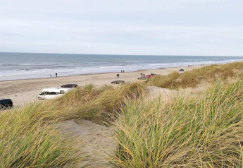 Vue depuis un lodge sur une plage avec des voitures, des dunes herbeuses et des gens près de l’océan.