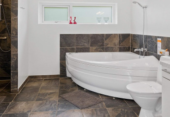Baño moderno con bañera de esquina, ducha, inodoro y azulejos oscuros en una casa de vacaciones en Blokhus, Dinamarca.