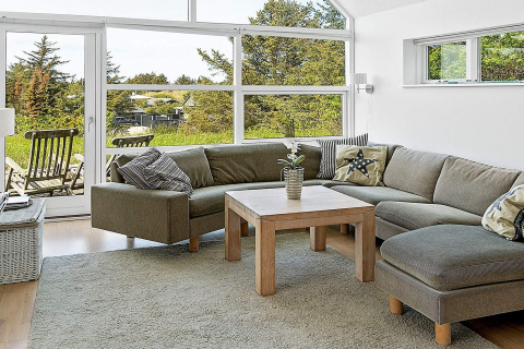 Helles Wohnzimmer mit Ecksofa, Holztisch und großen Fenstern mit Blick ins Grüne in einem Ferienhaus.