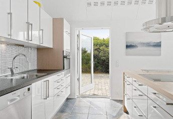 Cocina moderna en casa de vacaciones en Blokhus, Dinamarca, con gabinetes blancos y puerta abierta al jardín.
