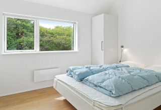 Slaapkamer met tweepersoonsbed, blauw dekbed, witte kast en groot raam met groene natuur in Blokhus.