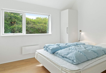 Chambre avec lit double, couette bleue, armoire blanche et grande fenêtre donnant sur la verdure à Blokhus.