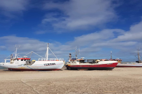 Fischerboote stehen am Sandstrand bei blauem Himmel und Wolken, vermutlich an einer Fischerhütte.