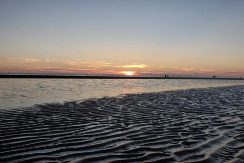 Sonnenuntergang am Strand bei einer Lodge mit seichtem Wasser, Sandwellen und Spaziergängern am Horizont.