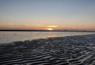 Sonnenuntergang am Strand bei einer Lodge mit seichtem Wasser, Sandwellen und Spaziergängern am Horizont.