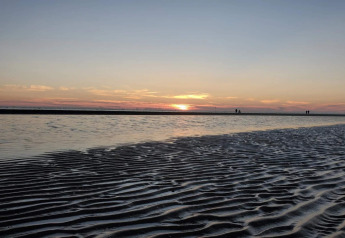 Sonnenuntergang am Strand bei einer Lodge mit seichtem Wasser, Sandwellen und Spaziergängern am Horizont.