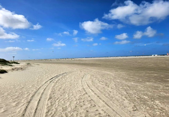 Bredt sandstrandområde med bilspor nær en lodge, blå himmel og skyer på en solrig dag.