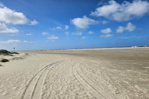 Weitläufiger Sandstrand mit Autospuren vor einer Lodge, blauer Himmel und wenige Wolken.