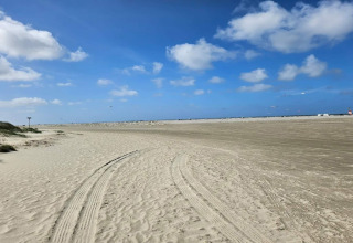 Weitläufiger Sandstrand mit Autospuren vor einer Lodge, blauer Himmel und wenige Wolken.