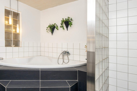 Helles Badezimmer in Seaside Charm in Rindby, Dänemark, mit Eckbadewanne, weißen Fliesen und Hängepflanzen.
