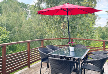 Terrasse am Ferienhaus in Gränna, Schweden, mit Glasstisch, vier Stühlen, rotem Sonnenschirm und Waldblick.