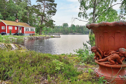 Casetta rossa sul lago ad Åkersberga, Svezia, con grande urna e vegetazione rigogliosa in primo piano.