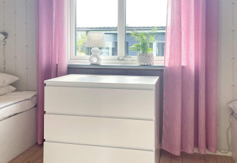 Chambre lumineuse avec commode blanche, rideaux roses, lampe et plante dans une maison de vacances à Gränna, Suède.