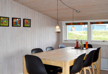 Comedor con mesa de madera, seis sillas negras, arte colorido en la pared y grandes ventanas en Fanø.