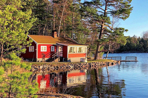 Casa vacanze rossa sul lago ad Åkersberga, Svezia, immersa tra pini e con un pontile sull’acqua.