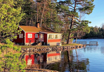 Rotes Ferienhaus in Åkersberga am Seeufer, umgeben von Kiefern, mit Steg in sonniger schwedischer Natur.