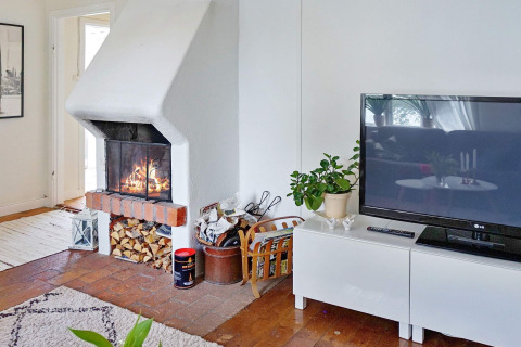 Wohnzimmer eines Ferienhauses in ÅKERSBERGA, Schweden, mit Kamin, Fernseher und Holzvorrat.