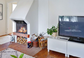 Wohnzimmer eines Ferienhauses in ÅKERSBERGA, Schweden, mit Kamin, Fernseher und Holzvorrat.