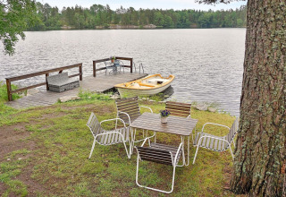 Sitzbereich im Freien mit Tisch und Stühlen am See, Steg und gelbes Ruderboot, umgeben von Wald.