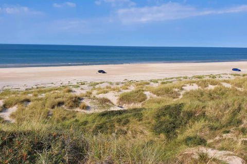 Udsigt fra glamping indkvartering over klitter og strand med to biler og roligt hav i baggrunden.