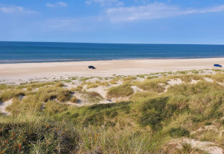 Blick von einer Glamping-Unterkunft auf sandige Dünen, den Strand, zwei Autos und das ruhige Meer.