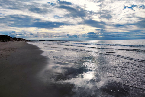 Fredelig strand med blødt skum, roligt hav og dramatiske skyer ved en glamping oplevelse ved kysten.