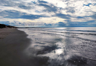 Fredelig strand med blødt skum, roligt hav og dramatiske skyer ved en glamping oplevelse ved kysten.