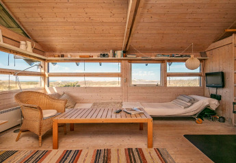 Indbydende stue i et sommerhus i Bindslev, Danmark, med lyst træinteriør, sofa og udsigt til naturen.
