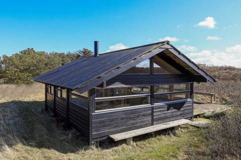 Schwarzes Holzchalet für 4 Personen in Bindslev, Dänemark, inmitten von Natur und unter blauem Himmel.