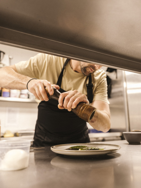 Un chef con delantal negro muele pimienta sobre un plato en la cocina de Wecamp Cala Montgó, Cataluña, España.