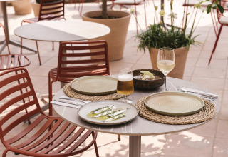Mesa de cafetería al aire libre con comida y bebidas junto a la piscina, rodeada de sillas rojas en Wecamp Cala Montgó.