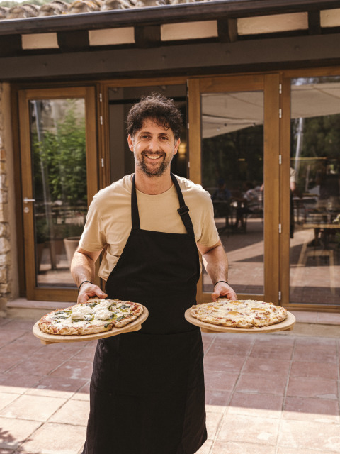 Mann im schwarzen Schürze und beigem T-Shirt serviert zwei Pizzen vor Wecamp Cala Montgó in Katalonien.